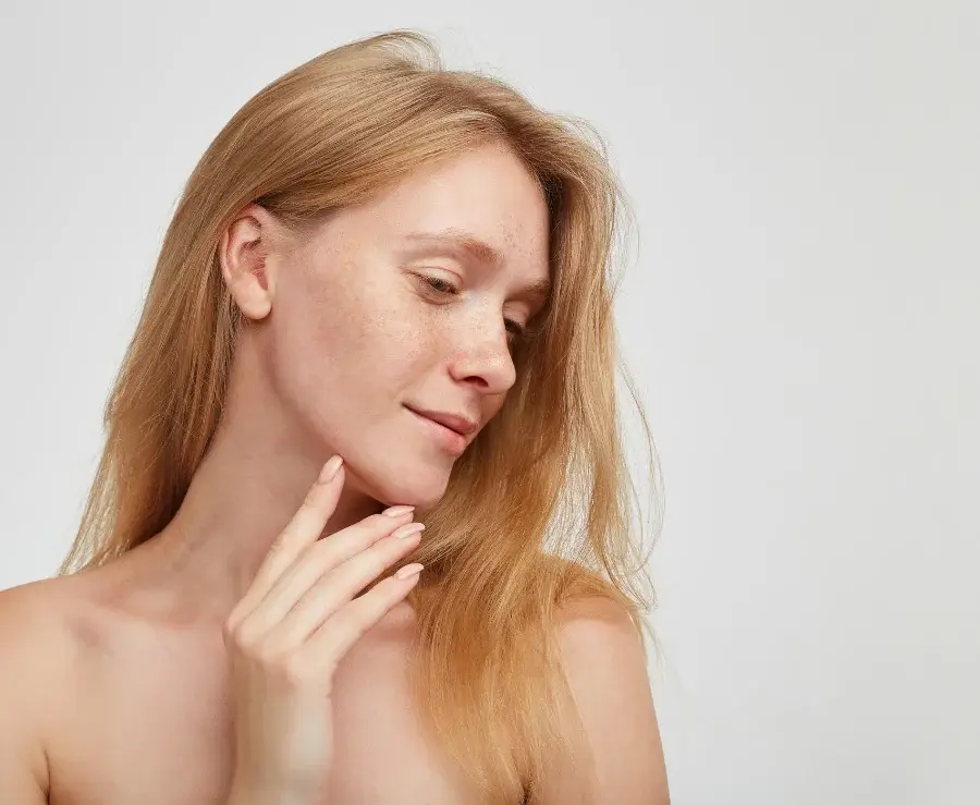 romantic-young-pretty-redhead-woman-with-long-hair-touching-her-chin-with-raised-hand-smiling-softly-standing-against-white-wall