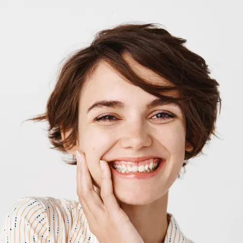 Close up of cute girl with short hair smiling with white teeth and touching natural clean face, standing over wall